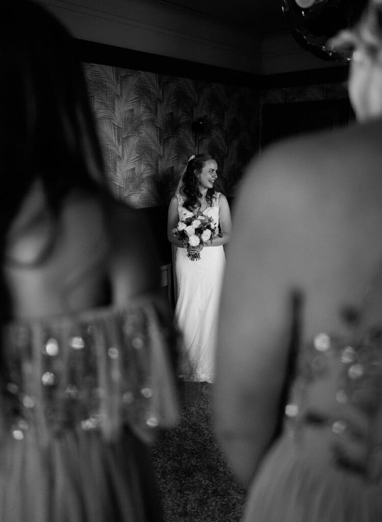 A bride reveals her dress to the bridesmaids, seen from their perspective