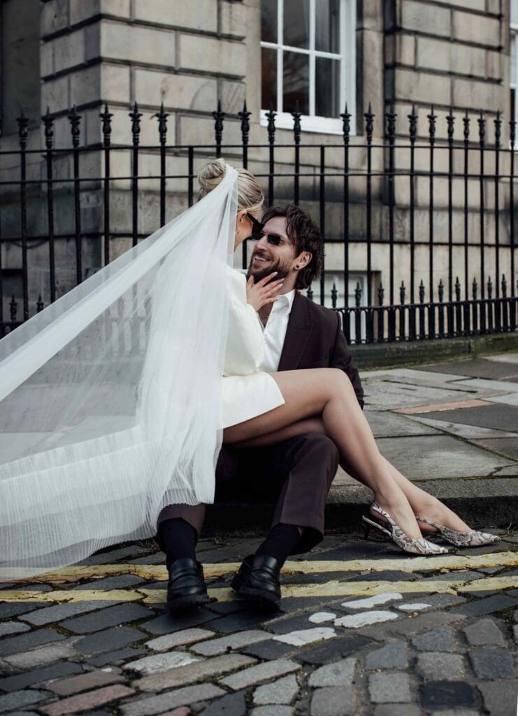 Bride sits on the grooms lap in a candid moment on the cobbled streets of Edinburghs west end
